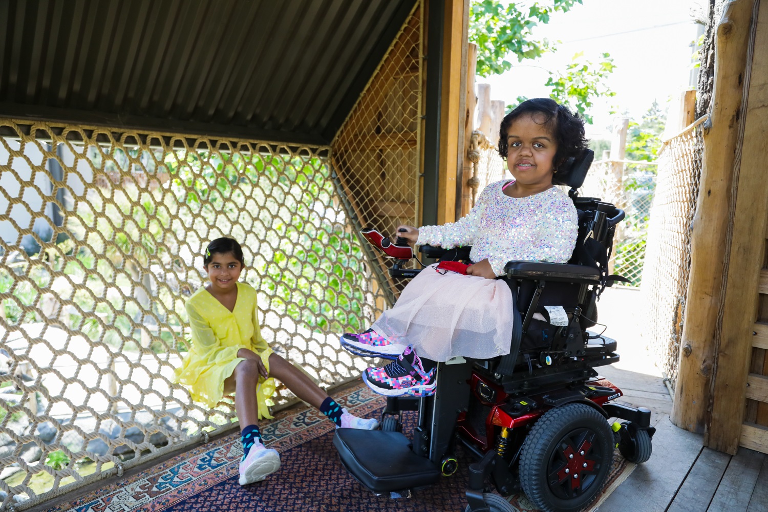 Two children sit side by side: one in a wheelchair, and one on the netting of the Tree House