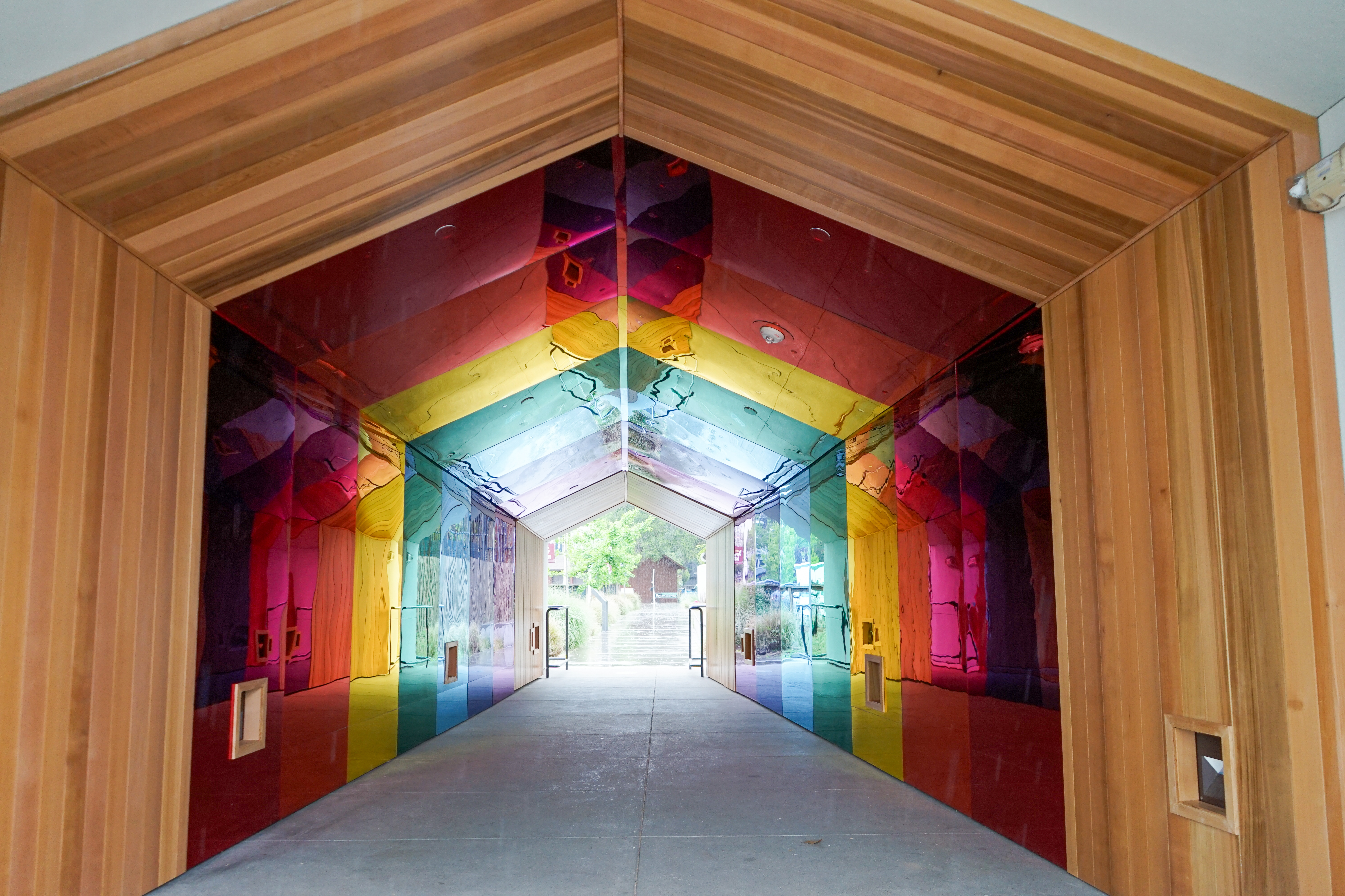 Entrance to a rainbow glass tunnel framed in wood, with mirrored colorful panels leading to an outdoor walkway.